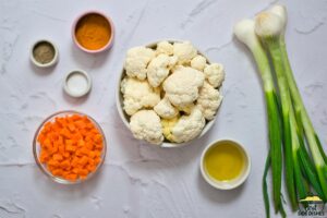 cauliflower rice ingredients on a white surface: spices, chopped carrots, cauliflower, olive oil, and onions