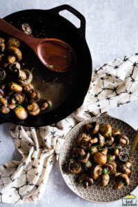 Button mushrooms in a cast iron skillet next to a plate of mushrooms