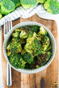 a bowl of cooked broccoli on a wooden board next to a fork and raw broccoli