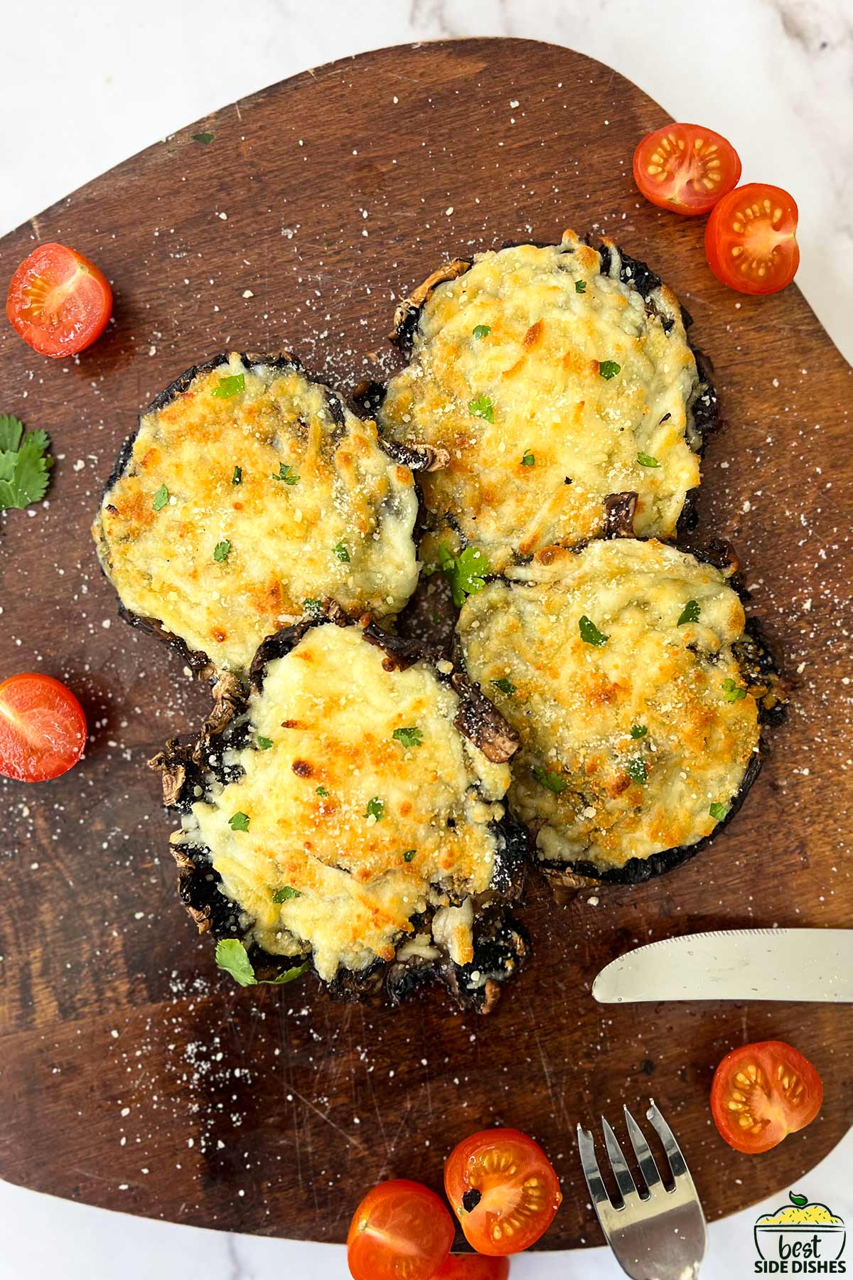 top down view of a stack of stuffed mushrooms on a cutting board with tomatoes and utensils