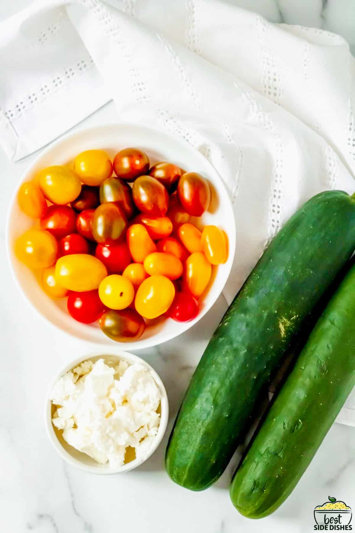cherry tomatoes in a bowl next to two cucumbers and feta in a bowl